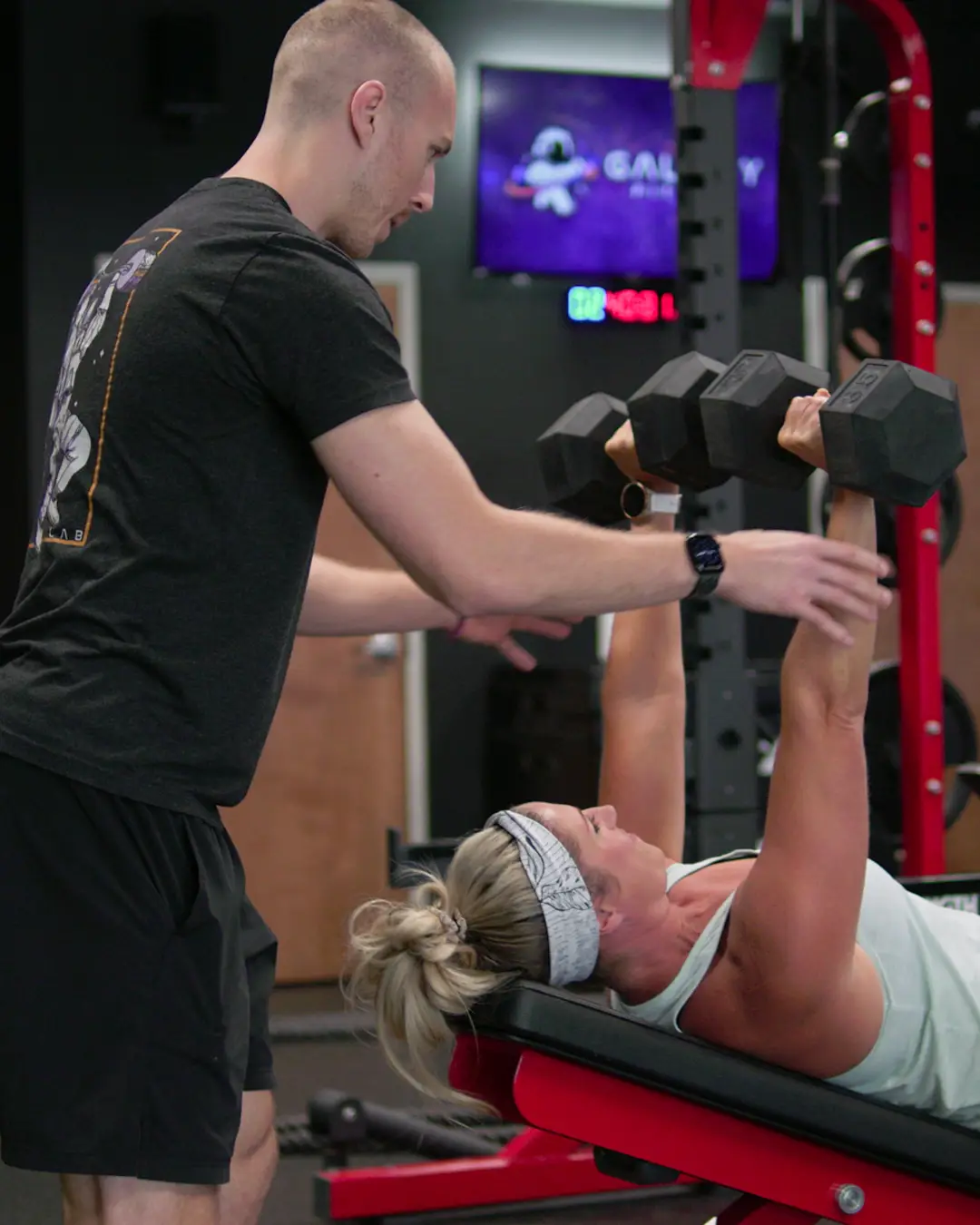 Personalized strength training with proper form - male trainer assisting female client with dumbbell bench press at Galaxy Fit Lab Naples